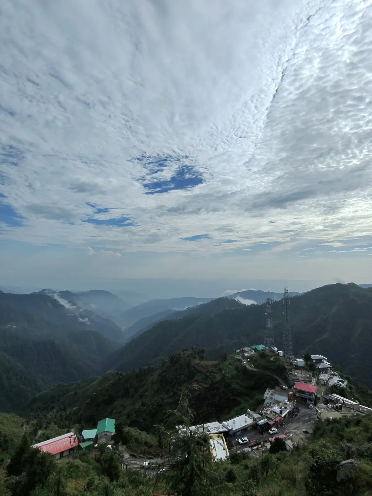 Suwakholi Gaon on its ridge near Mussoorie, with telecom towers, colourful roofs and forested valleys under a sky of clouds